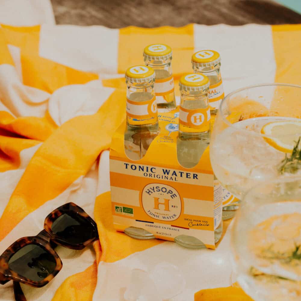 Pack de 4 tonics pour cocktails de la marque Hysope sur une serviette de plage jaune, avec des lunettes de soleil et des verres à cocktail