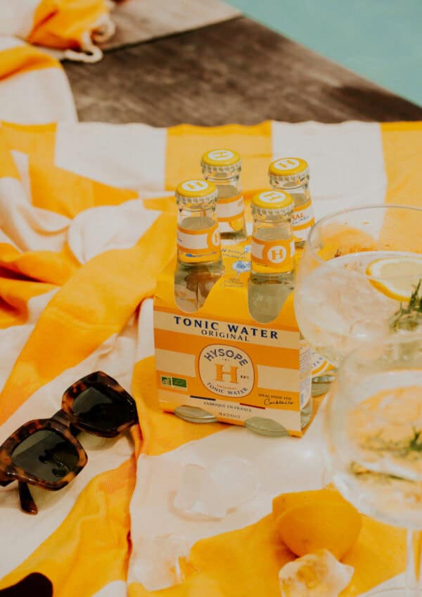 Pack de 4 tonics pour cocktails de la marque Hysope sur une serviette de plage jaune, avec des lunettes de soleil et des verres à cocktail