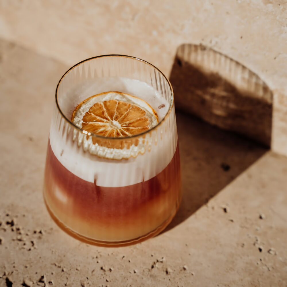 Cocktail réalisé lors d’un atelier cocktail à Aix-Les-Bains avec Chasarose. Cours de mixologie. Un cocktail avec une mousse blanche, une tranche d'orange séchée et une couleur orange.